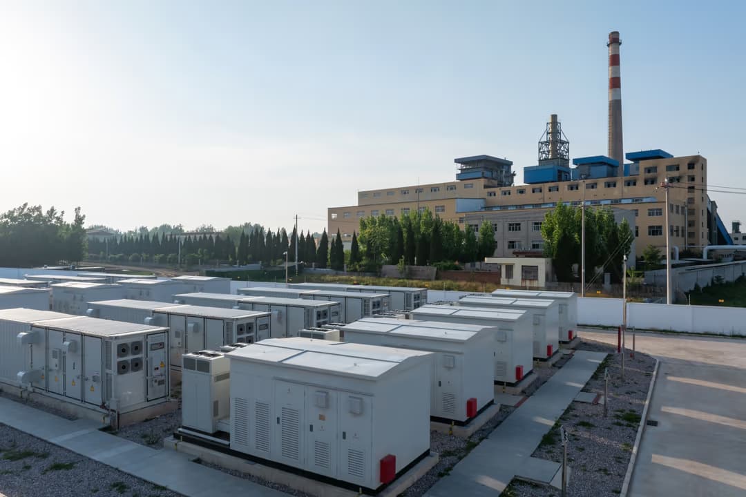 Battery energy storage cabinet arrays installed at an industrial facility with a factory building in the background, representing the physical infrastructure that enables on-site peak shaving, demand charge reduction, and grid services participation for commercial and industrial operations