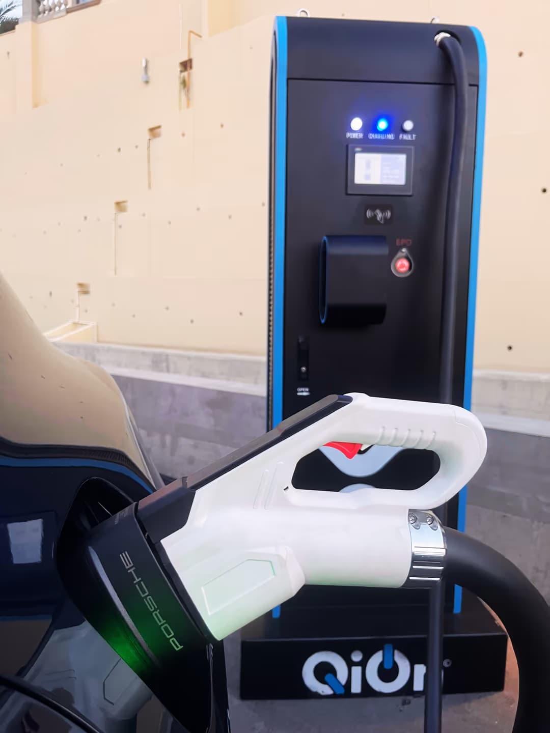Close-up of a Porsche-branded CCS charging connector plugged into the Cayenne Electric, with the QiOn Maveriq supercharger showing an illuminated charging status light behind it