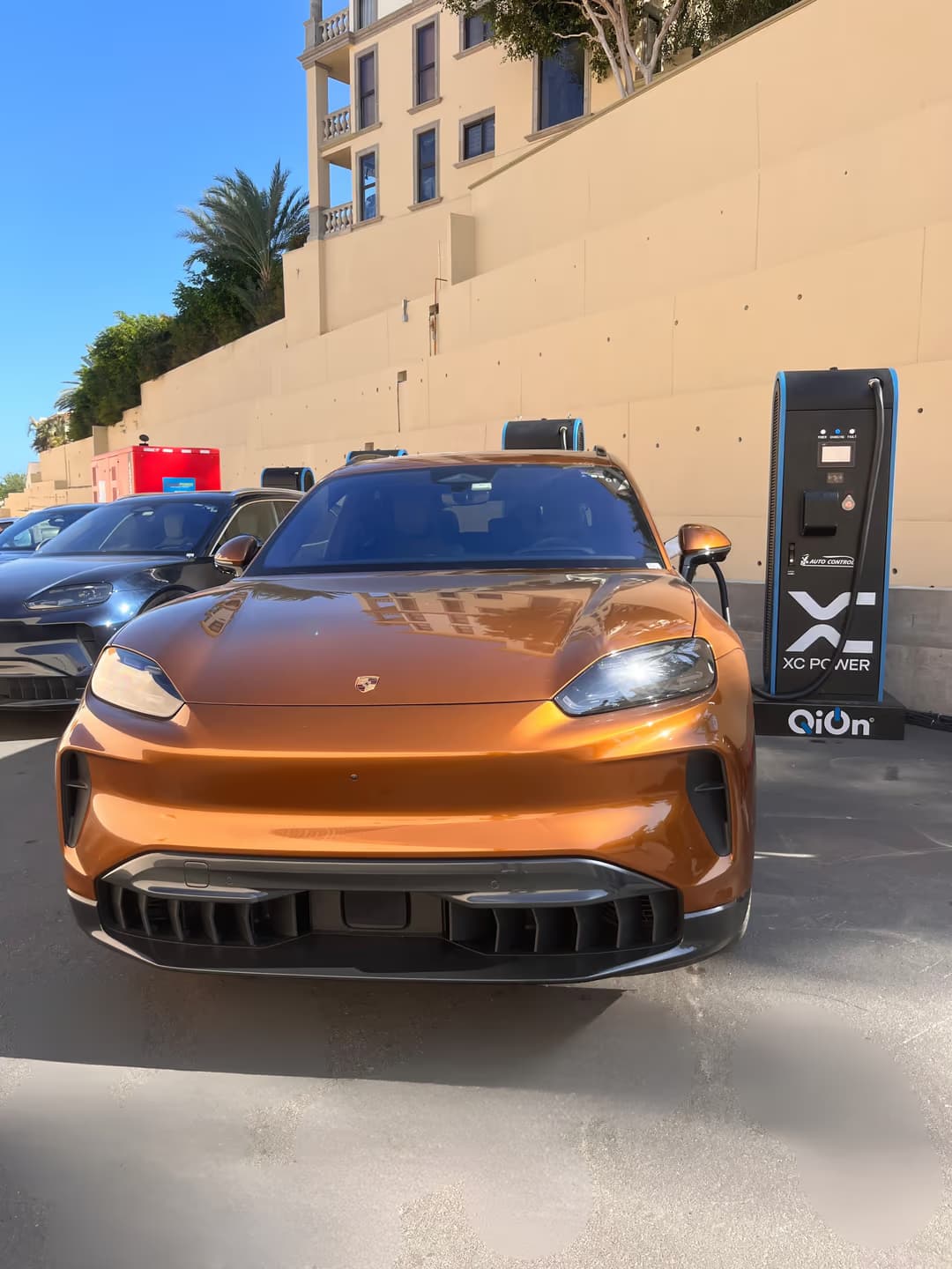 An orange Porsche Cayenne Electric plugged into a QiOn Maveriq DC supercharger at the Los Cabos event, with additional Cayenne vehicles and the off-grid power unit in the background
